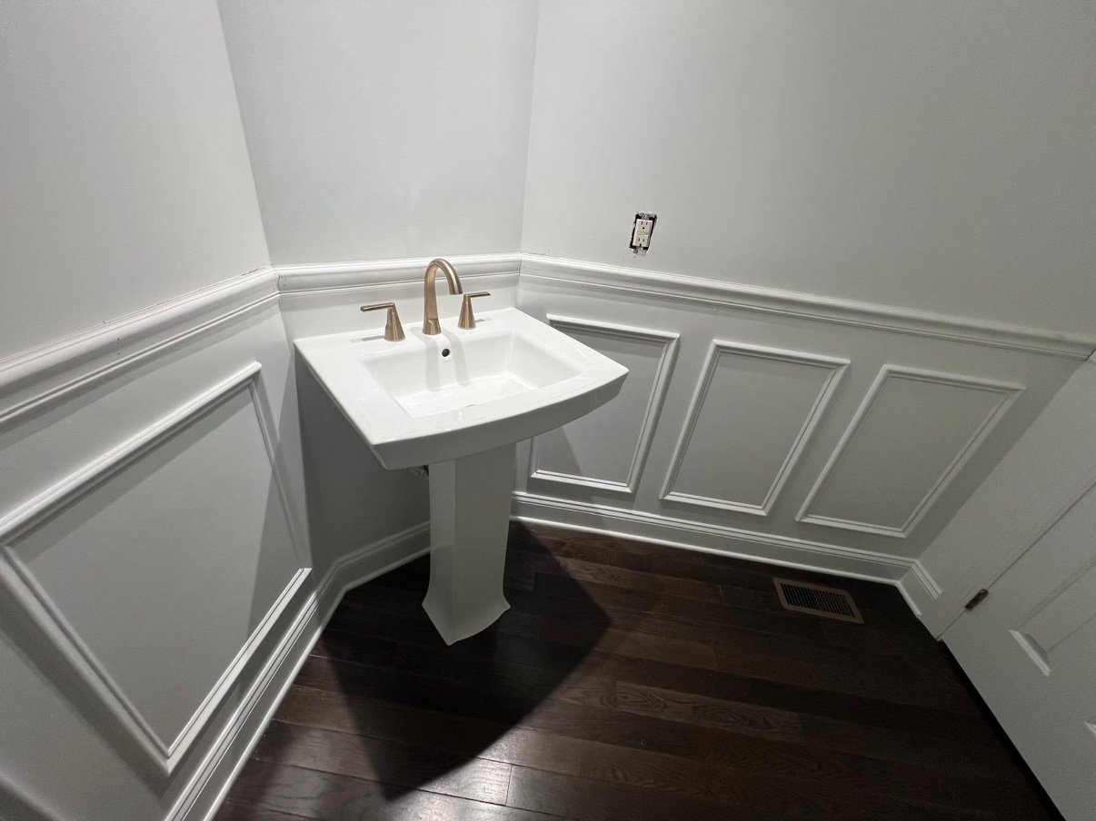 Powder room with a pedestal sink, white wainscoting paneling, dark wood floor, and a brushed gold faucet
