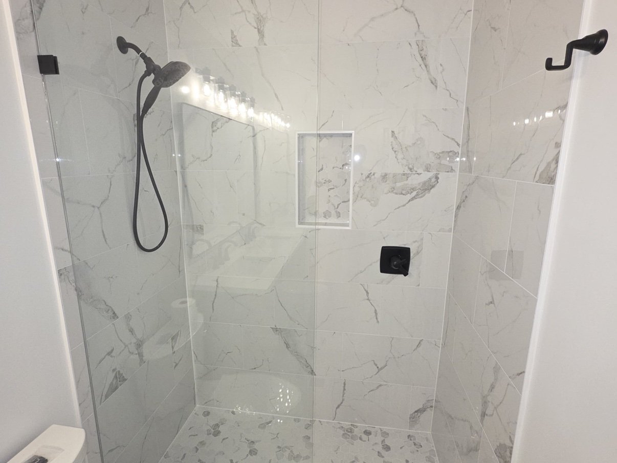 White marble-look tile walk-in shower with frameless glass panel, hexagon mosaic floor, and matte black fixtures, remodeled by Carolina Construction Bros